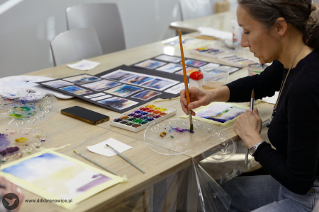 Uczestniczka w czarnej bluzce maluje obraz. Pędzel moczy w rozlanej na plastiku wodzie wymieszanej z farbą.