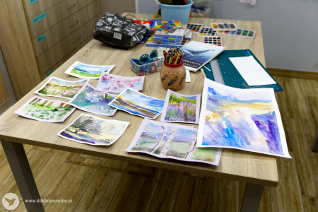 Gotowe obrazy. Przedstawiają krajobrazy. Obok nich leżą bloki papieru i pędzle.