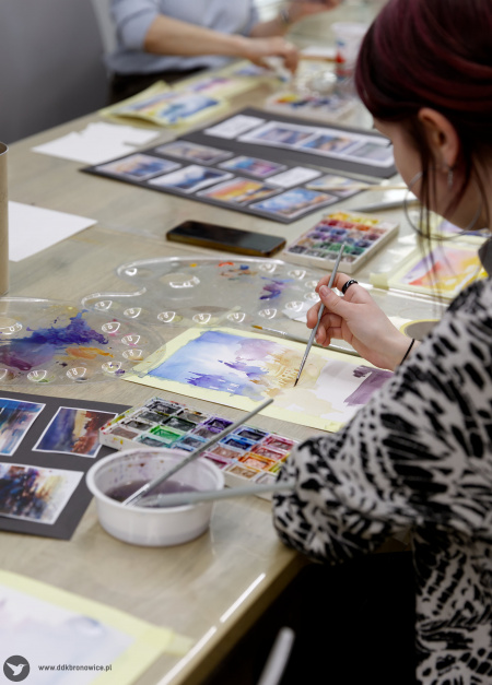 Uczestniczka maluje swój obraz. Obok niej na stole są farby i kubeczek z wodą.
