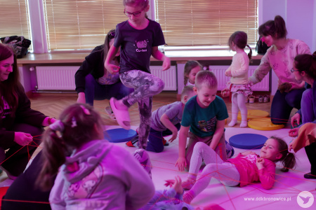 Dzieci bawią się czerwoną wstążką na białej macie. Dzieci śmieją się. Zapalone jest fioletowe światło.