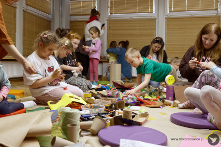 Dzieci tworzą artystyczne ozdoby. Na macie leżą włóczki, nożyczki i artykuły papiernicze. Mamy pomagają swoim dzieciom.