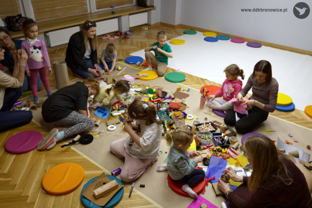 Dzieci i rodzice tworzą ozdoby artystyczne na macie. Uczestnicy siedzą na kolorowych poduszkach.