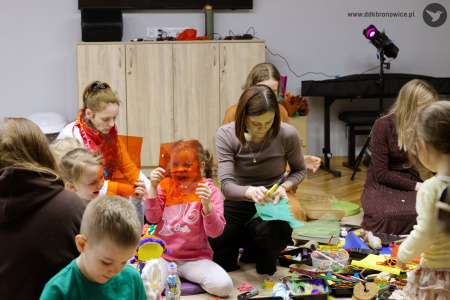 Dzieci tworzą ozdoby artystyczne. Dziewczynka przyłożyła do swojej twarzy przezroczysty pomarańczowy kartonik. Śmieje się.