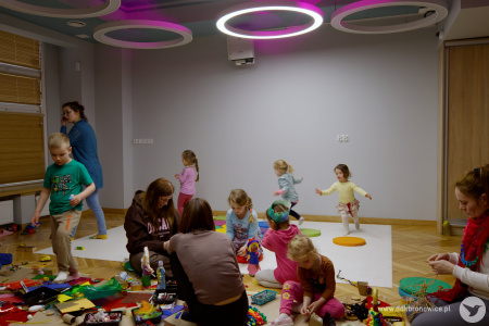 Grupa dziewczynek biega po białej macie. Z przodu osoby pracują nad swoimi ozdobami.