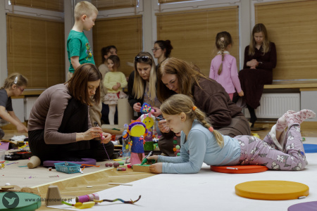 Uczestnicy warsztatów pracują nad ozdobami. Dziewczynka w błękitnej bluzce leży na małej pomarańczowej poduszce.