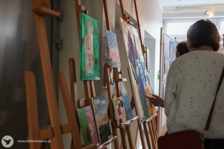 Na sztalugach wyłożone mniejsze obrazy. Widać osoby, które wskazują na elementy obrazów palcami.