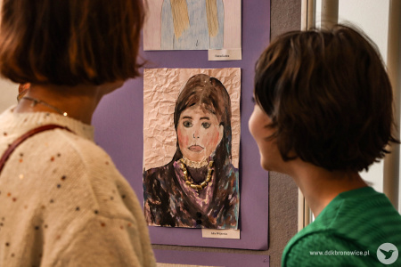 Dwie osoby oglądają wystawę malarską dzieci. Między nimi portret kobiety w czarnej sukni.