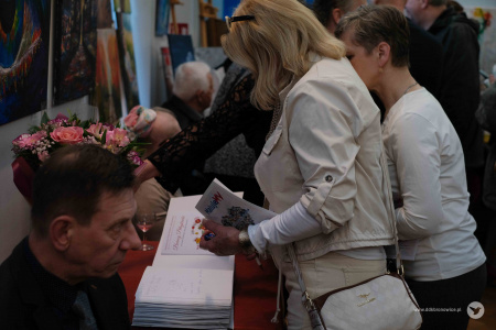 Dwie uczestniczki wystawy oglądają przy stole foldery dotyczące wystawy. Obok stolika siedzi mężczyzna w czarnej marynarce.