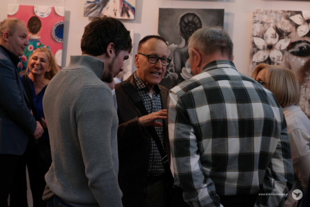 Trzech mężczyzn rozmawia w trakcie wystawy. Mężcyzna po prawej ubrany jest w koszulę w kratkę, mężczyzna po lewej w szary sweter, a mężczyzna w stojący w środku w ciemną koszulę i marynarkę.