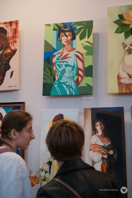 Dwie kobiety oglądają obrazy wiszące na ścianie. Jeden z nich przedstawia kobietę o krótkich włosach, na tle geometrycznych i florystycznych figur.