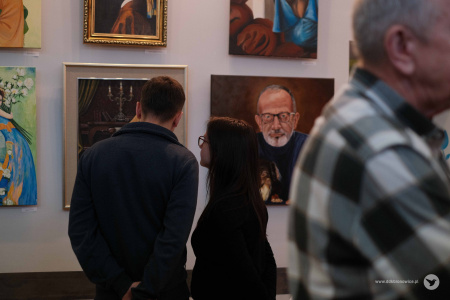 Mężczyzna w czarnym swetrze i kobieta w czarnej bluzce oglądają wystawę obrazów. Jeden z obrazów przedstawia starszego mężczyznę z krótką, siwą brodą i w okularach.