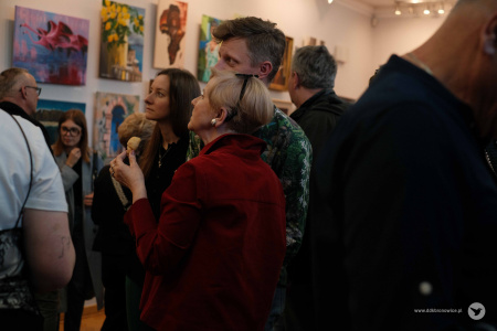 Kobieta w bordowym żakiecie rozmawia z mężczyzną. Kobieta trzyma w ręce rogalika. Zwróceni są w kierunku obrazów.