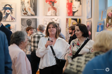 Dwie kobiety w jasnych koszulach rozmawiają. Mają przewieszone torby. Kobieta po lewej stronie trzyma kieliszek.