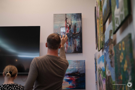 Mężczyzna w szarym swetrze robi zdjęcie telefonem obrazowi. Obraz jest impresjonistyczny i przedstawia statek.