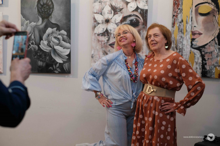 Prowadząca grupę, Monika Szydłowska, pozuje do zdjęcia z kobietą ubraną w brązową sukienkę na tle zawieszonych obrazów.