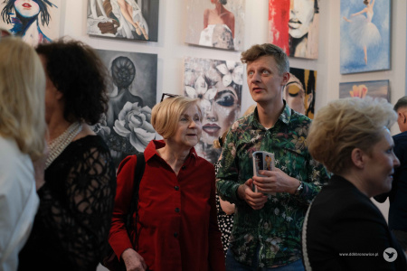Kobieta w czerwonej koszuli i mężczyzna w koszuli w kwiaty rozmawiają. W tle widać obrazy i osoby oglądające wystawę.