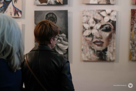 Kobieta w czarnej skórze ogląda zawieszony obraz. Obraz przedstawia twarz kobiety z zamkniętymi oczami. Na oku widać duży kwiat.