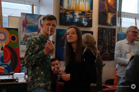 Mężczyzna w koszuli w kwiaty rozmawia z kobietą. Wskazuje zwiniętą kartką w kierunku wystawy.