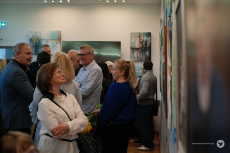 Po prawej stronie zawieszone obrazy. Po lewej stronie uczestnicy wystawy.