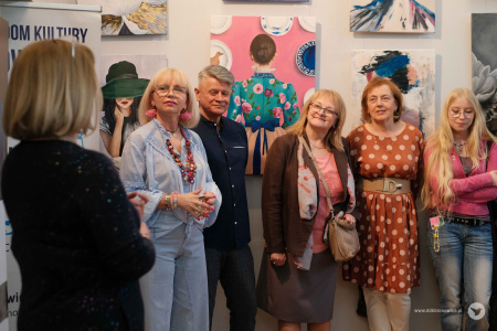 Uczestnicy grupy MalujeMY stoją na tle obrazów. Po lewej stronie kobieta ubrana w czarną sukienkę.