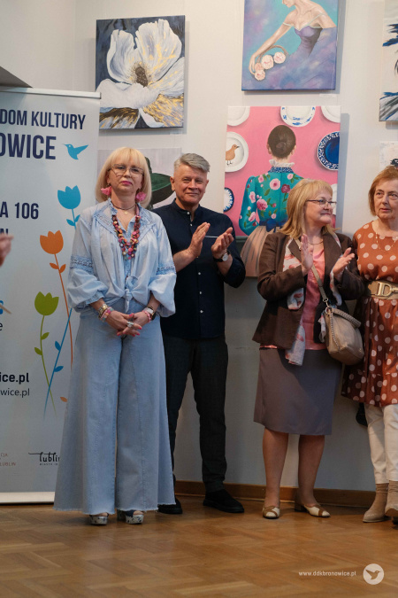 Monika Szydłowska stoi obok uczestników grupy. Za osobami wiszą duże obrazy.