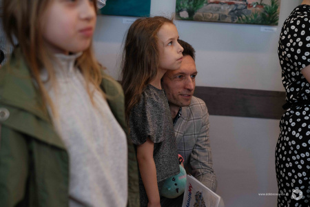 Mężczyzna kuca obok małej dziewczynki. Dziewczynka patrzy w kierunku obrazów. Ubrana jest w szarą koszulkę.