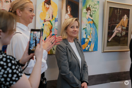 Na tle zawieszonych obrazów stoi Ewa Rejmak. Po lewej stronie kobieta robi zdjęcie telefonem obrazom.