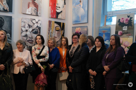 Osoby zgromadzone na wernisażu. Za nimi duże okno. Obok okna, na ścianie, zawieszone duże obrazy.