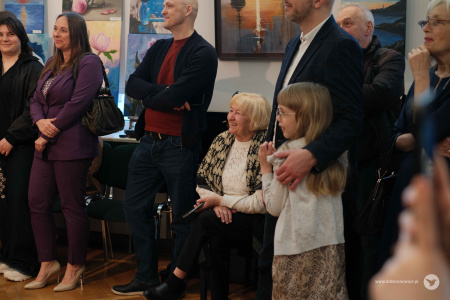Osoby zgromadzone na wernisażu słuchają przemówień. Starsza kobieta siedzi. Obok niej stoją mężczyźni. Mężczyzna po prawej stronie przytula małą dziewczynkę.