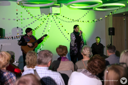 Na pierwszym planie widownia. W tle wokalistka śpiewa do mikrofonu po prawej stronie. Po lewej stoi gitarzysta. Za nimi zielone światło.