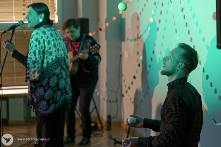 Po prawej stronie artysta żongluje. Po lewej stronie śpiewa wokalistka. W tle gitarzysta gra na gitarze.