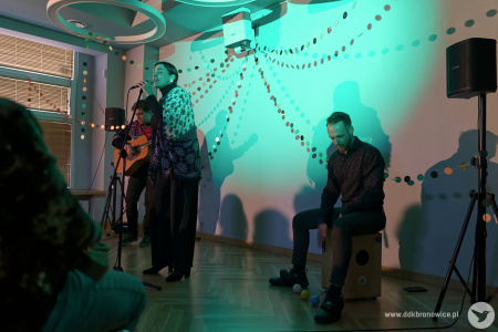 Artyści grają koncert. Po prawej stronie mężczyzna gra na cajonie. Pośrodku wokalistka śpiewa. W tle gitarzysta. Artystów oświetla błękitne światło.