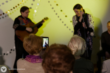 W tle na muzyków pada żółte światło. Na pierwszym planie, kobieta na widowni robi zdjęcie telefonem.