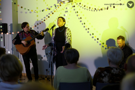 Wokalistka zespołu śpiewa emocjonalną piosenkę. Ma zamknięte oczy. Oświetla artystkę żółte światło.