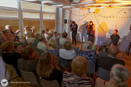 Na artystów pada pomarańczowe światło. Muzycy grają piosenkę. Na pierwszym planie siedząca widownia.