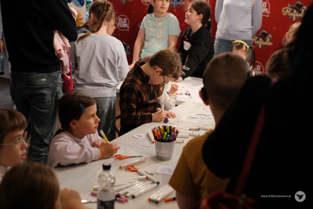 Dzieci kolorują fragmenty papierowej ozdoby. Na środku stoi chłopiec w koszuli w kratę.
