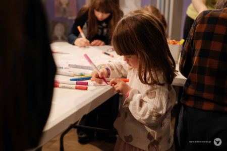 Dziewczynka w białym swetrze w misie koloruje fragment swojej ozdoby. Na stole obok niej leżą flamastry.