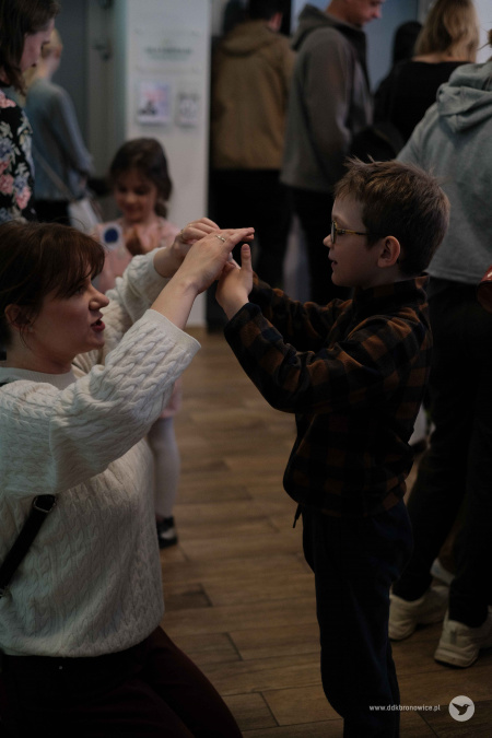 Mama w szarym swetrze trzyma chłopca za ręce. Mają je uniesione w górze. Chłopiec uśmiecha się.