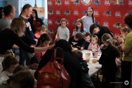 Przy stole bardzo dużo grupa dzieci tworzy papierowe ozdoby. Wśród nich stoją dorośli. W tle widać na czerwonym tle logotyp Magic Show.