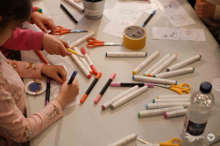 Na białym stole widać rozrzucone flamastry, wodę w butelce, nożyczki, taśmę klejącą i niewycięte fragmenty ozdób na białych kartkach.