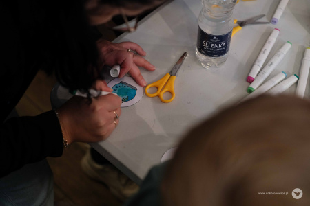 Osoba kolorują fragment ozdoby. Przedstawia okrągłe akwarium. Na stole stoi butelka wody i żółte nożyczki.