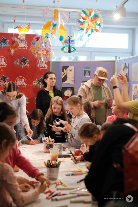Dzieci wciąż pracują nad papierowymi ozdobami. Dorośli im pomagają. Z sufitu zwisają kolorowe ozdoby o kształtach owadów.