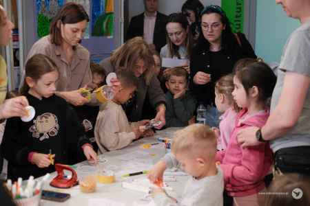 Dzieci i dorośli wspólnie tworzą ozdoby. Dzieci są najbliżej stołu. Dorośli stoją nad nimi.
