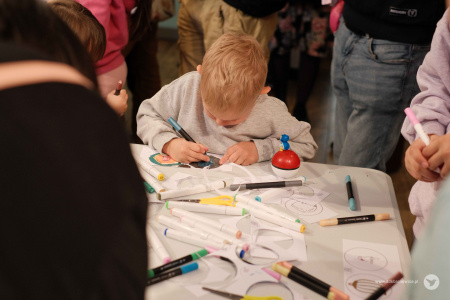 Chłopiec w jasnej bluzie koloruje ozdobę. Na stole rozłożone flamastry i nożyczki. Chłopiec jest bardzo skupiony.