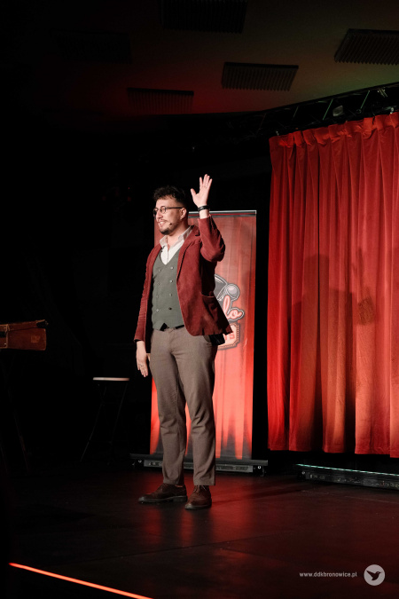 Na scenie w czerwonej marynarce macha magik do publiczności. Jest bardzo uśmiechnięty.