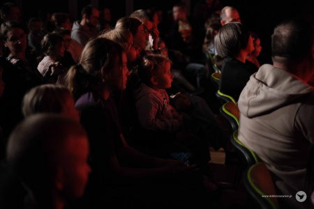 Osoby siedzące na widowni. Oświetlone są delikatnym czerwonym światłem. Oglądają występ magiczny.