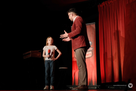 Magik w czerwonej marynarce rozmawia z dziewczynką w białej koszulce na scenie. Dziewczynka trzyma mikrofon.
