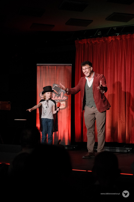 Dziewczynka i Magik stoją obok siebie na scenie. Dziewczynka wskazuje palcem na widownie. Magik bardzo wyraźnie śmieje się do widowni.