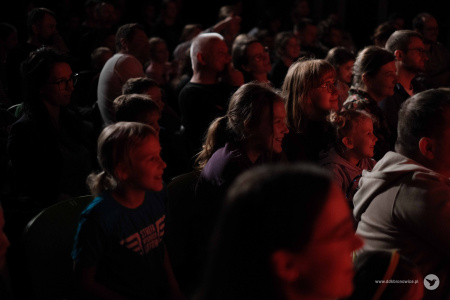 Osoby na widowni szeroko uśmiechają się. Oświetleni czerwonym światłem oglądają występ magiczny.