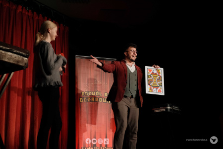 Magik wita na scenie dziewczynę w szarym swetrze. Dziewczyna trzyma mikrofon. Magik pokazuje dużą kartę: dama czerwo.